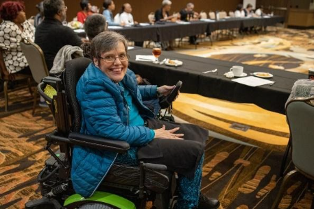 A woman in a wheelchair smiles at the camera, while sitting at a large table during a meeting.